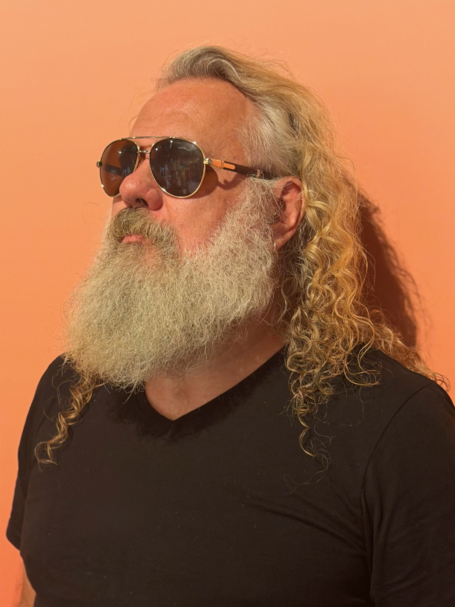 Gaz Williams with sunglasses, long hair and beard against a warm orange background
